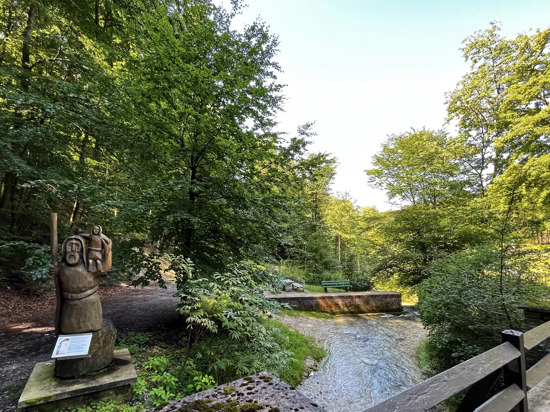 Foto Christophorus Statue.jpeg Eine hölzerne Christophorus-Statue steht am Waldesrand nahe einem sprudelnden Bach in grüner Umgebung.