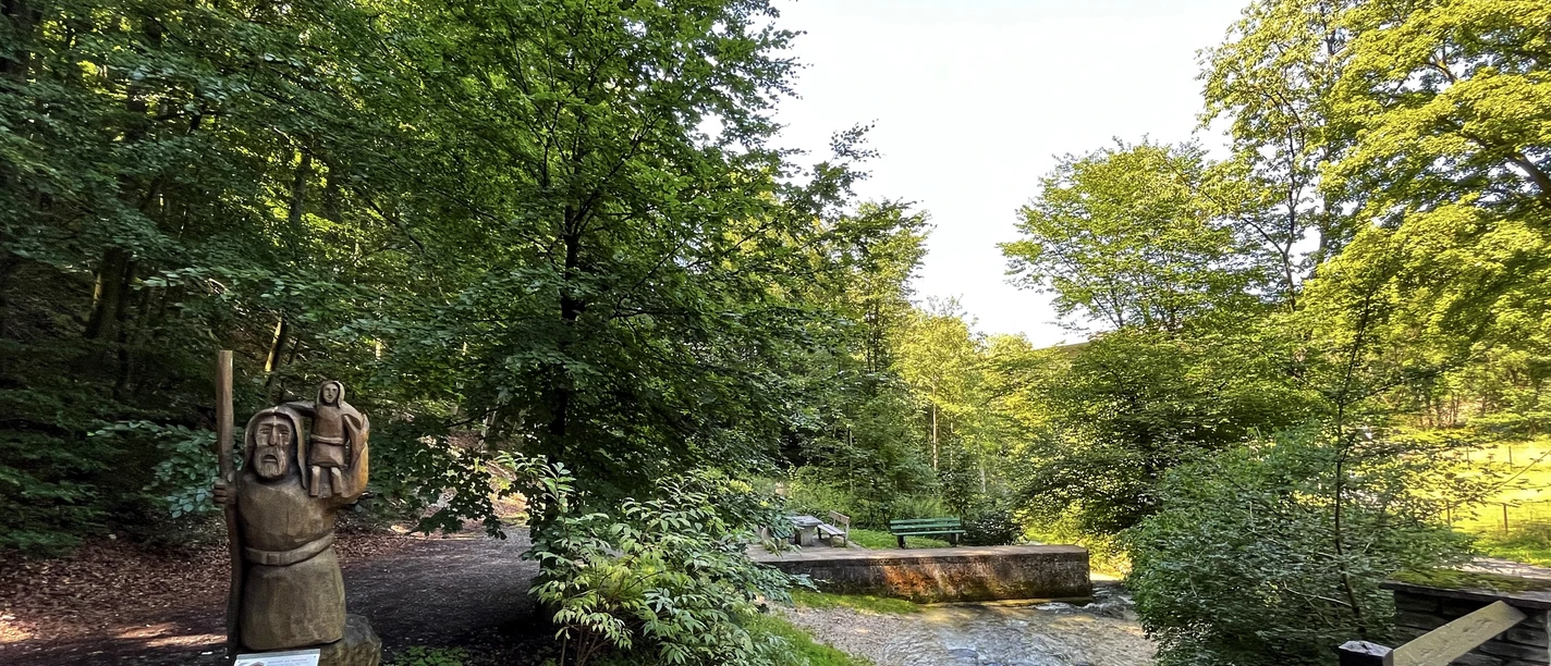 Foto Christophorus Statue.jpeg Eine hölzerne Christophorus-Statue steht am Waldesrand nahe einem sprudelnden Bach in grüner Umgebung.
