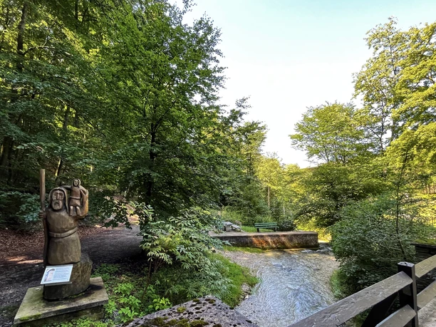 Foto Christophorus Statue.jpeg Eine hölzerne Christophorus-Statue steht am Waldesrand nahe einem sprudelnden Bach in grüner Umgebung.