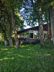 Foto Bauernkamp.jpeg Die Holzhütte mit großer Fensterfront ist von Bäumen umgeben und steht am Waldrand in grüner Umgebung.