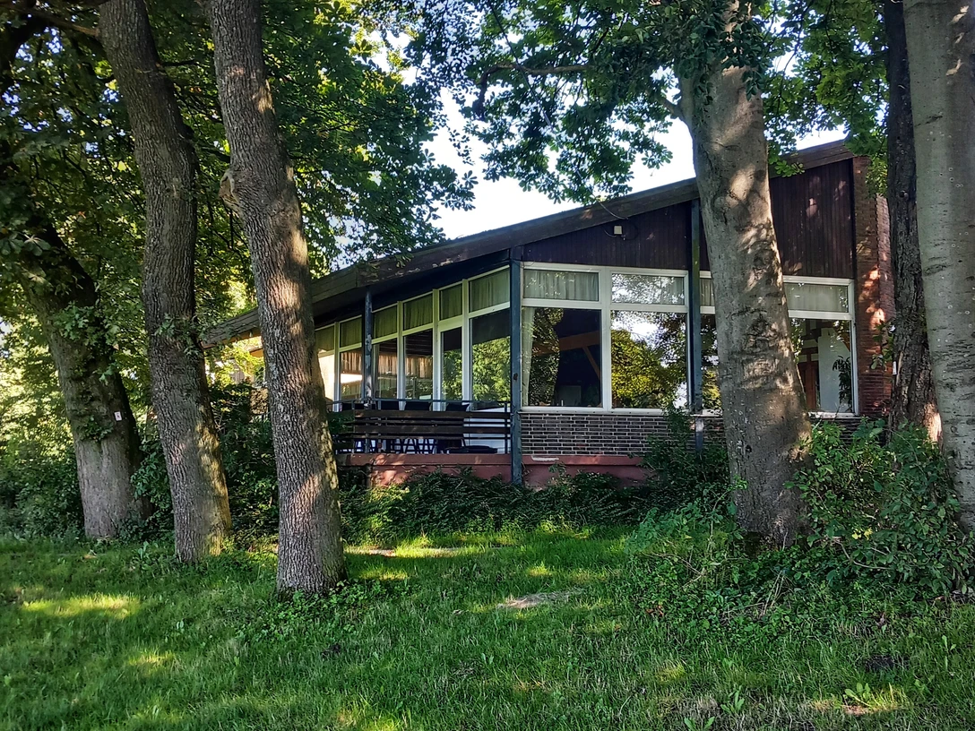 Foto Bauernkamp.jpeg Die Holzhütte mit großer Fensterfront ist von Bäumen umgeben und steht am Waldrand in grüner Umgebung.