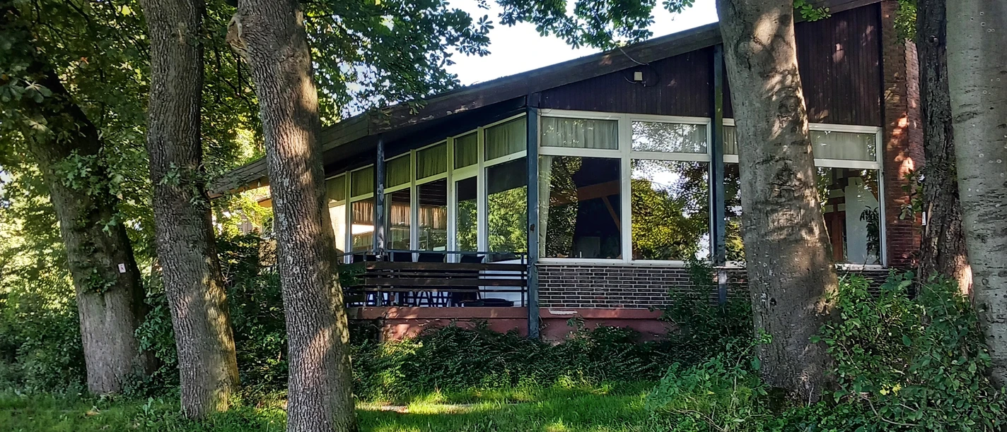 Foto Bauernkamp.jpeg Die Holzhütte mit großer Fensterfront ist von Bäumen umgeben und steht am Waldrand in grüner Umgebung.