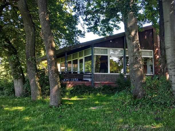 Foto Bauernkamp.jpeg Die Holzhütte mit großer Fensterfront ist von Bäumen umgeben und steht am Waldrand in grüner Umgebung.
