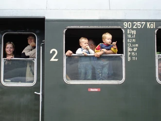 Mediendatei Menschen blicken aus den Fenstern eines historischen grünen Zugwaggons, darunter zwei kleine Kinder.
