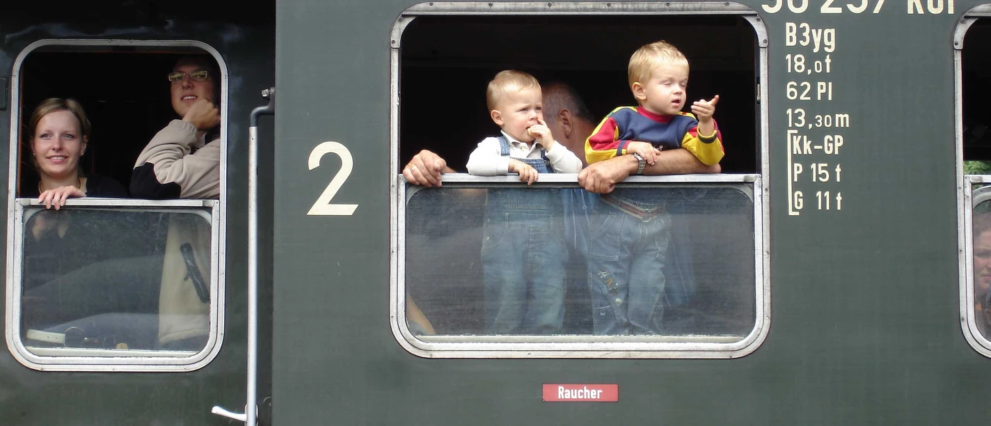 Mediendatei Menschen blicken aus den Fenstern eines historischen grünen Zugwaggons, darunter zwei kleine Kinder.
