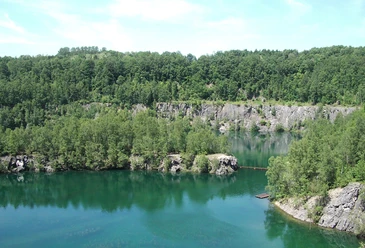 Steinbruch Schlupkothen Azurblauer See, umgeben von üppigen Wäldern und Felsklippen unter klarem, sonnigem Himmel.