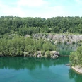 Steinbruch Schlupkothen Azurblauer See, umgeben von üppigen Wäldern und Felsklippen unter klarem, sonnigem Himmel.