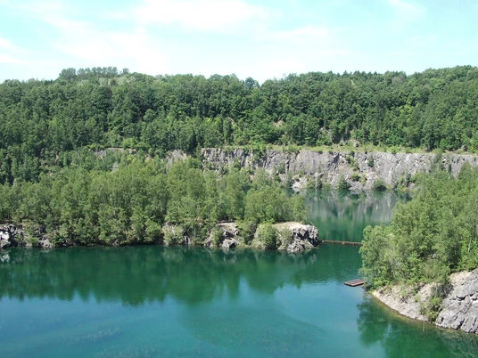 Steinbruch Schlupkothen Azurblauer See, umgeben von üppigen Wäldern und Felsklippen unter klarem, sonnigem Himmel.