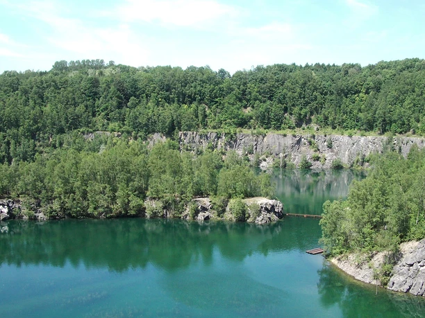Steinbruch Schlupkothen Azurblauer See, umgeben von üppigen Wäldern und Felsklippen unter klarem, sonnigem Himmel.