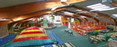 Mediendatei Indoor-Spielplatz mit bunten Hüpfburgen, Klettergerüsten und Sitzbereichen unter Holzdecken.