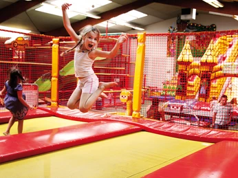 Mediendatei Ein Mädchen springt freudig auf einem roten Trampolin in einer bunten Indoor-Spielhalle.