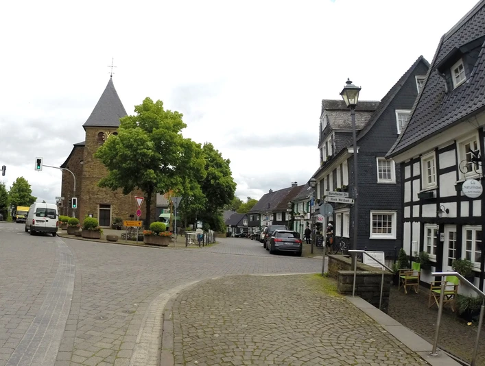Historischer Ortskern Witzhelden Dorfstraße mit historischem Fachwerkhaus und Kirche im Hintergrund, umgeben von grünen Bäumen.