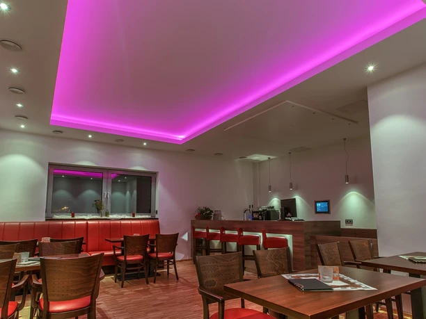 life-ness Innenbereich Gemütlicher Speiseraum mit lila beleuchtetem Deckenelement, roten Sitzbänken und Holztischen.