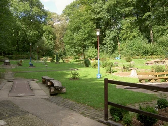 Minigolfplatz Ein gepflegter Minigolfplatz inmitten eines grünen Parks, umgeben von hohen Bäumen und Sitzbänken.