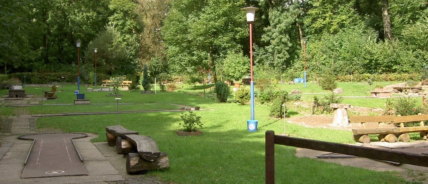 Minigolfplatz Ein gepflegter Minigolfplatz inmitten eines grünen Parks, umgeben von hohen Bäumen und Sitzbänken.