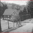 Mediendatei Schwarzweiß-Foto eines traditionellen Waldbauernhauses mit steilem Satteldach am Rande eines Waldwegs.