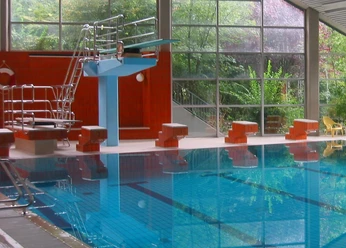 Walter-Leo-Schmitz-Bad Schwimmhalle mit großem Glasfenster, Sprungturm, roten Wänden und blauem, klarem Wasser.