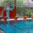 Walter-Leo-Schmitz-Bad Schwimmhalle mit großem Glasfenster, Sprungturm, roten Wänden und blauem, klarem Wasser.