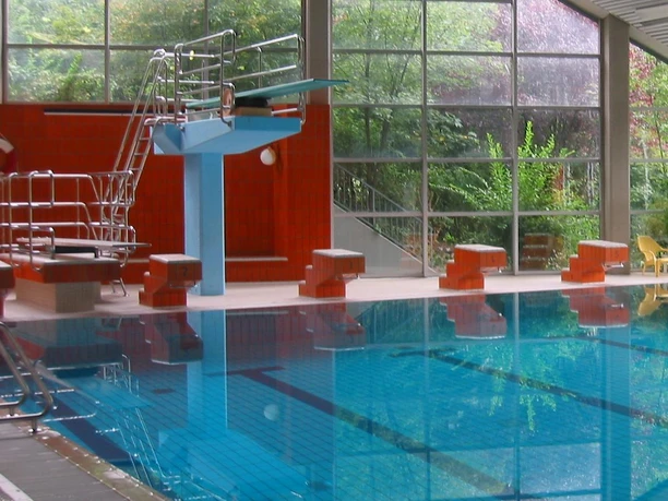 Walter-Leo-Schmitz-Bad Schwimmhalle mit großem Glasfenster, Sprungturm, roten Wänden und blauem, klarem Wasser.