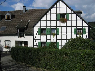 Mediendatei Fachwerkhaus mit grünen Fensterläden, weißen Wänden und Dachgauben, umgeben von Hecken bei Sonnenschein.