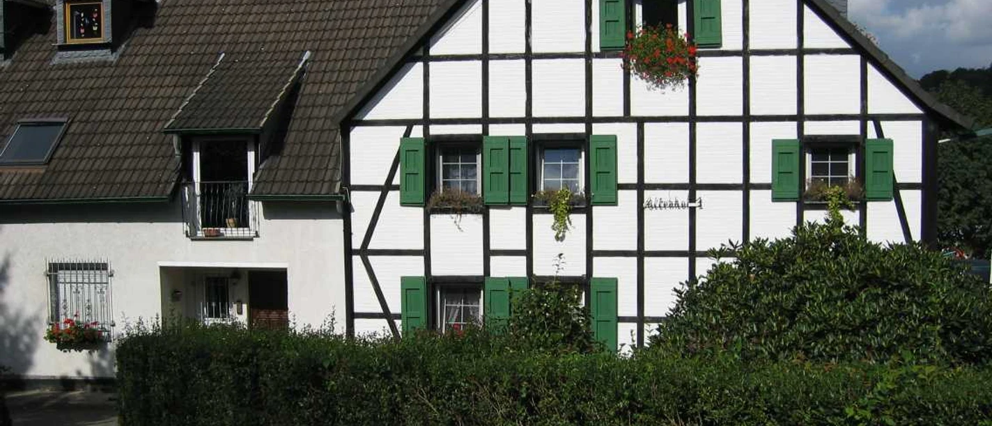 Mediendatei Fachwerkhaus mit grünen Fensterläden, weißen Wänden und Dachgauben, umgeben von Hecken bei Sonnenschein.