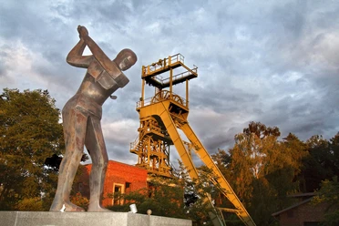 Golfclub Lüderich mit altem Förderturm Industriedenkmal mit gelbem Förderturm, flankiert von einer Bergarbeiterstatue und umgeben von Bäumen.