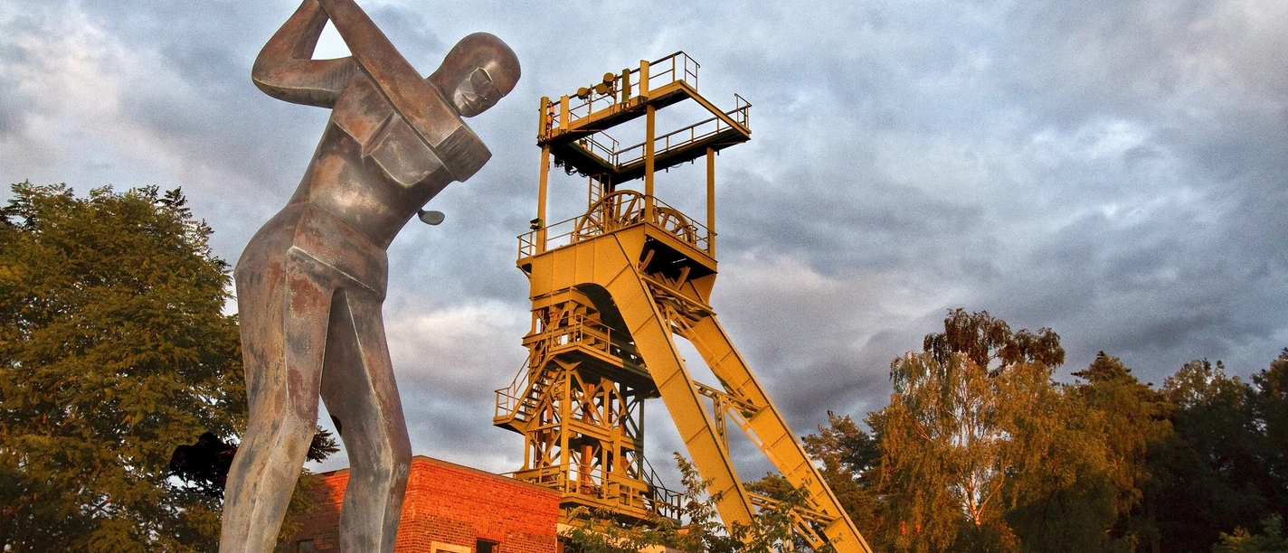Golfclub Lüderich mit altem Förderturm Industriedenkmal mit gelbem Förderturm, flankiert von einer Bergarbeiterstatue und umgeben von Bäumen.