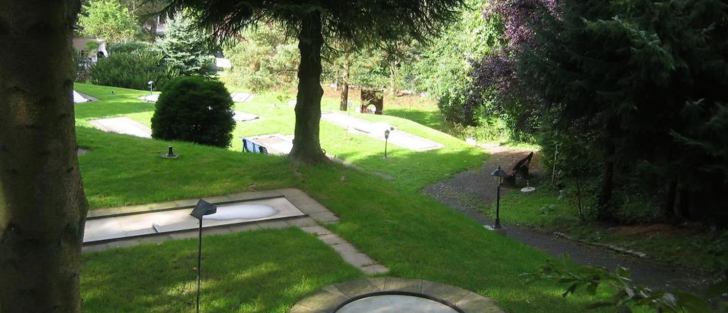 Mediendatei Eine Minigolfanlage im schattigen Wald, von Bäumen umgeben, mit gepflegtem Rasen.