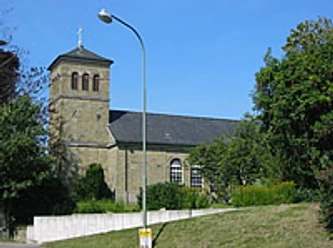 Mediendatei Romanische Steinkirche mit Satteldach und Glockenturm, umgeben von Grün und blauem Himmel.