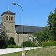 Mediendatei Romanische Steinkirche mit Satteldach und Glockenturm, umgeben von Grün und blauem Himmel.