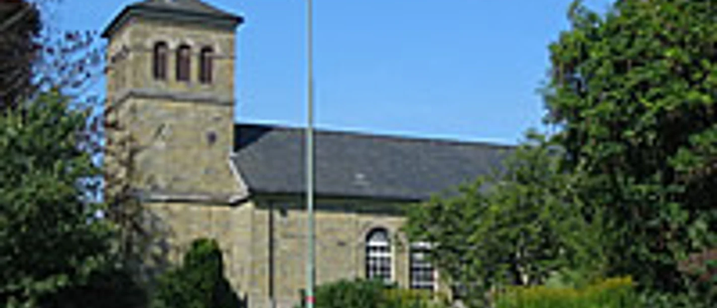 Mediendatei Romanische Steinkirche mit Satteldach und Glockenturm, umgeben von Grün und blauem Himmel.