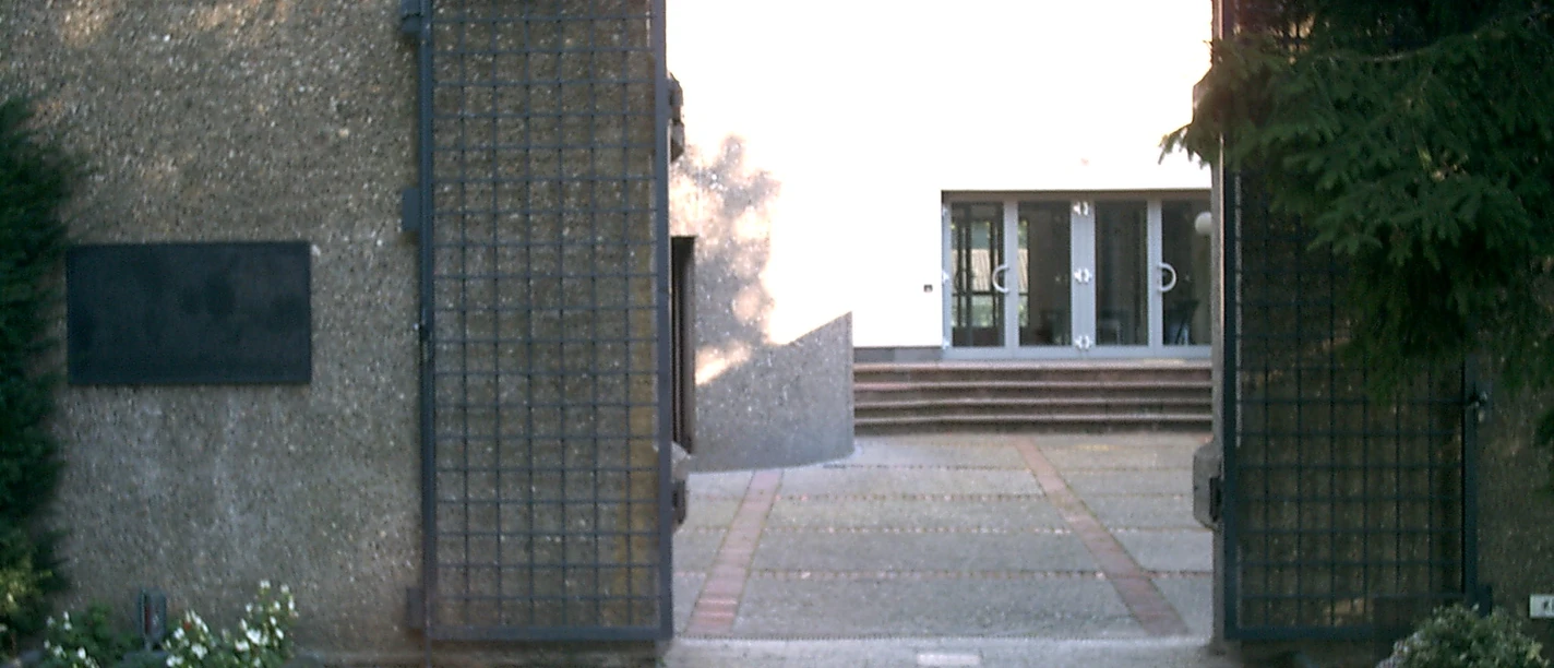 Mediendatei Geöffnete massive Eisentore führen zu einem Gebäude mit breiten Stufen, umgeben von grünen Bäumen.