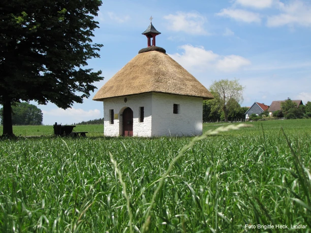 Kapelle "Frauenhäuschen" Kleines, strohgedecktes Kapellchen auf grüner Wiese, flankiert von Bäumen, blauer Himmel im Hintergrund.
