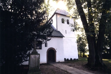 Mediendatei Kleine weiße Kirche mit Glockenturm, umgeben von Bäumen, mit einem alten Grabstein im Vordergrund.