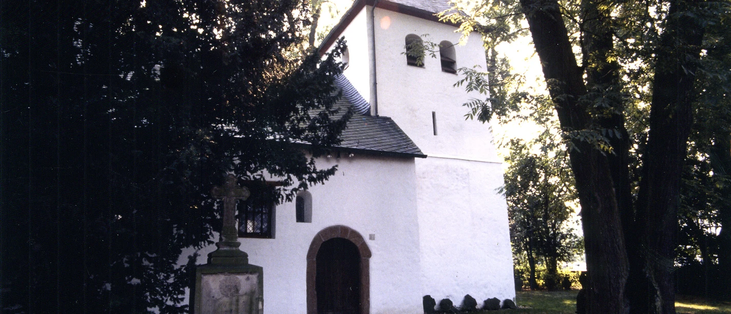 Mediendatei Kleine weiße Kirche mit Glockenturm, umgeben von Bäumen, mit einem alten Grabstein im Vordergrund.