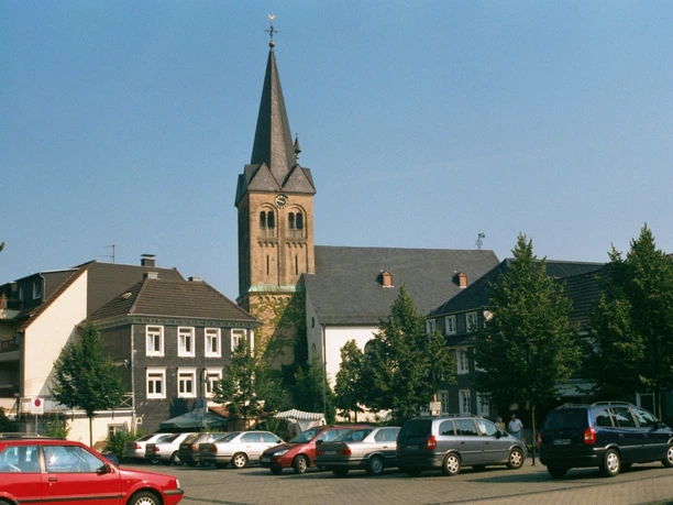Ev. Kirche Burscheid Kirche mit markantem Turm umgeben von historischen Fachwerkhäusern auf einem sonnigen Platz.
