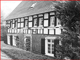 Grünscheider Mühle Fachwerkhaus mit weißen Wandflächen und schwarzen Holzbalken, umgeben von Bäumen und Sträuchern.