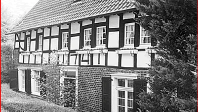 Grünscheider Mühle Fachwerkhaus mit weißen Wandflächen und schwarzen Holzbalken, umgeben von Bäumen und Sträuchern.