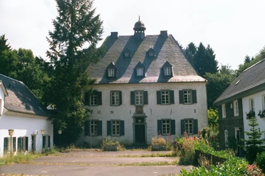Mediendatei Ein historisches Herrenhaus mit grauer Fassade und dunklem Dach, umgeben von Bäumen und Pflanzen.