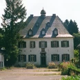Mediendatei Ein historisches Herrenhaus mit grauer Fassade und dunklem Dach, umgeben von Bäumen und Pflanzen.
