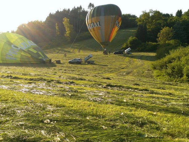 Ballonstartplatz Zwei bunte Heißluftballons auf einer offenen Wiese, umgeben von sanftem, grünem Hügelwald.