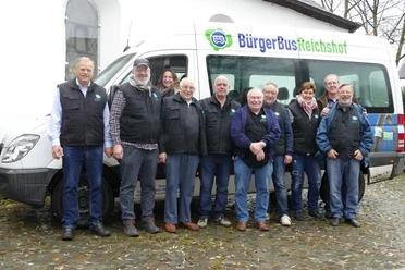 BürgerBus Verein Reichshof Elf Menschen posieren lächelnd vor einem Bürgerbus von Reichshof auf einem gepflasterten Platz.