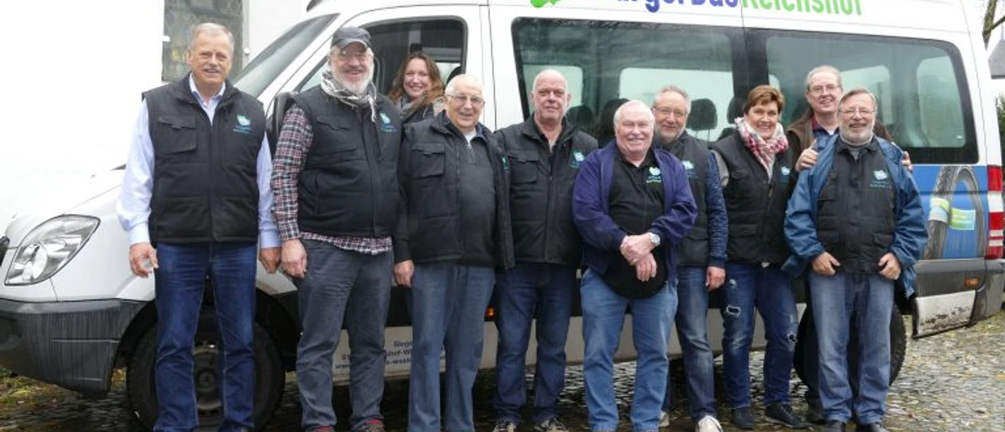 BürgerBus Verein Reichshof Elf Menschen posieren lächelnd vor einem Bürgerbus von Reichshof auf einem gepflasterten Platz.