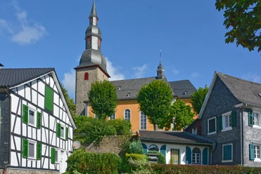 Ortskern von Eckenhagen mit ev. Barockkirche Fachwerkhäuser und eine Kirche unter blauem Himmel, umgeben von grünen Bäumen und Gehwegen.
