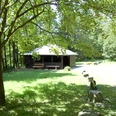 Grillhütte Windfus Ein hölzerner Pavillon steht in einem sonnigen Waldstück. Ein Pfad führt durch die grüne Landschaft.