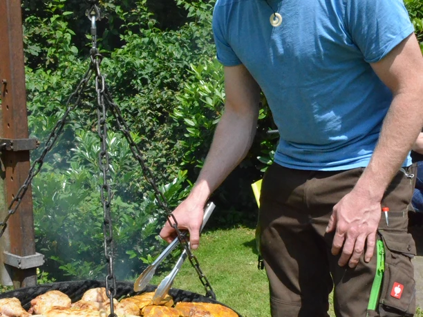 Grillen im Affen- und Vogelpark Ein Mann grillt Fleisch und Gemüse auf einem runden Rost im Freien, umgeben von grünem Laub.