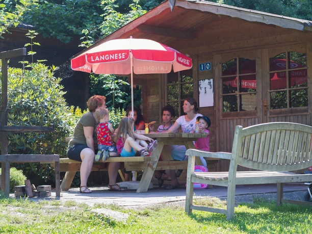 Grillhütte im Affen- und Vogelpark Familie und Freunde sitzen unter einem Sonnenschirm an einem Holztisch vor einer rustikalen Hütte.