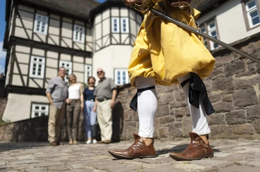 hx_fuehrung_jostziegenhirt_andreashub.jpg Eine Person in historischer Kleidung mit Schwert steht vor einer Gruppe vor Fachwerkhäusern.