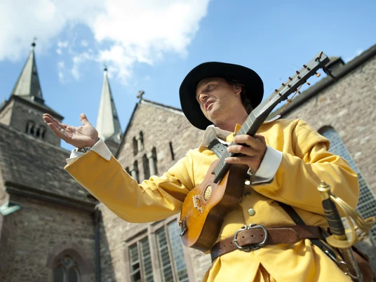 hx-jostziegenhirt-andreashub (4).jpg Ein mittelalterlich gekleideter Mann spielt Laute vor einer historischen Kirche an einem sonnigen Tag.