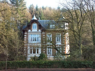 Villa Karl Reusch Charmante Villa mit weißem Dekor, umgeben von herbstlichen Bäumen und üppigem Grün, vor Waldkulisse.